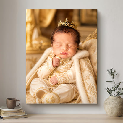 Newborn baby in royal attire with a crown, sitting on an ornate chair, framed on a wall.
